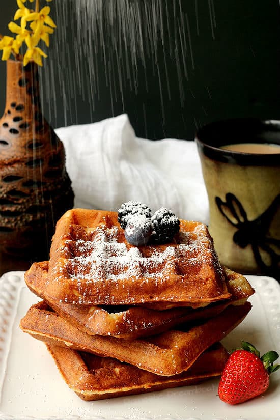Overnight Yeast-Raised Belgian Waffles on a white plate topped with berries being sprinkled with powdered sugar.