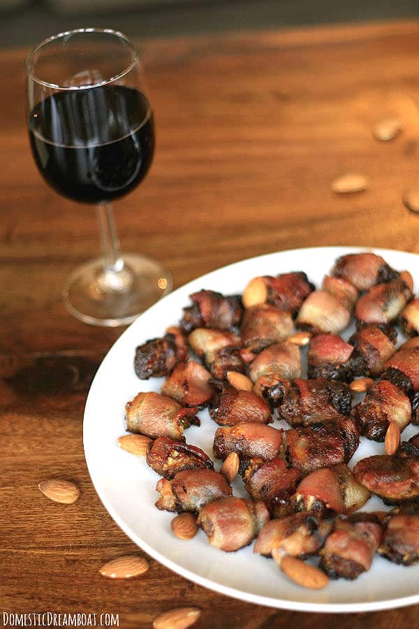 Bacon-Wrapped Stuffed Dates on a white plate with a glass of wine in the background.