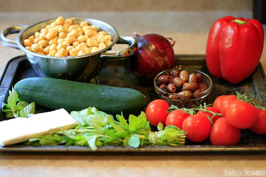 The ingredients to make chickpea and quinoa salad on a baking sheet: chickpeas, red onion, red pepper, cucumber, kalamata olives, tomatoes, oregano, Feta