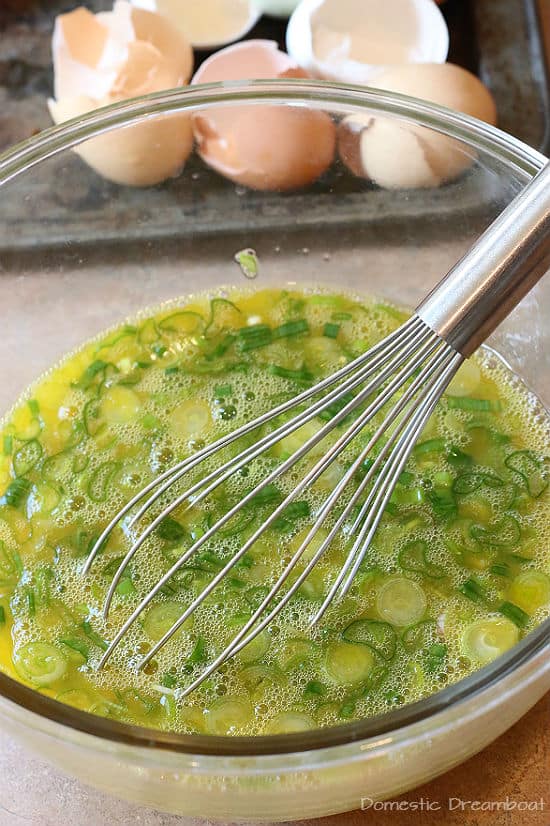 Raw eggs whisked together with sliced green onions in a glass bowl.