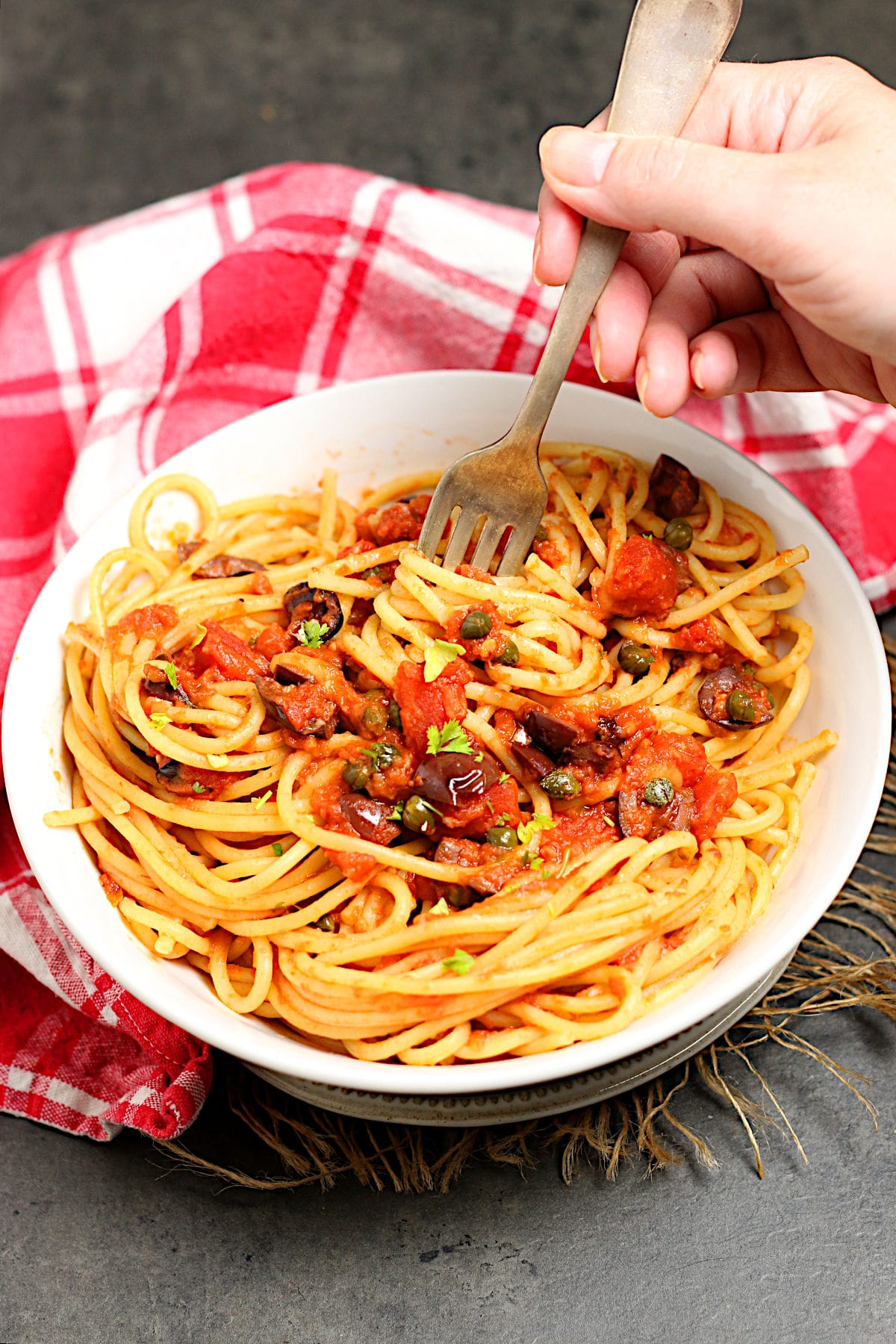 A fork scooping some spaghetti puttanesca from a white bowl.
