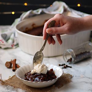 A hand placing a dollop of whipped cream on a bowl of gingerbread pudding cake.