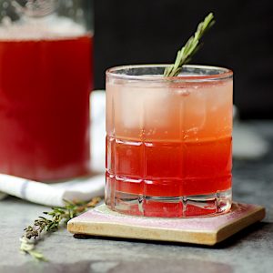 Strawberry shrub in a glass with ice, garnished with a sprig of rosemary.