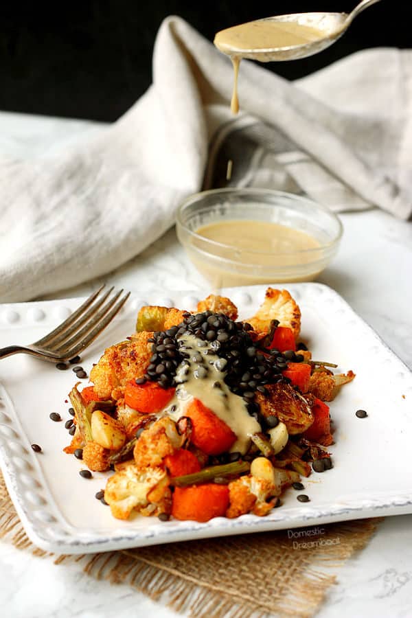 A spoon drizzing tahini dressing onto a plate of roasted vegetables and black lentils