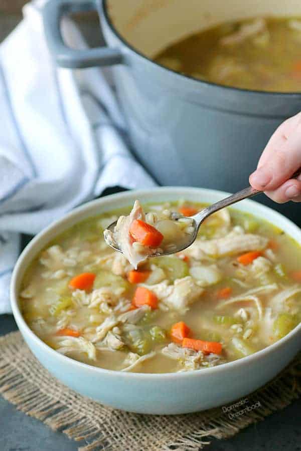 A spoon of chicken barley soup held over a bowl of soup.