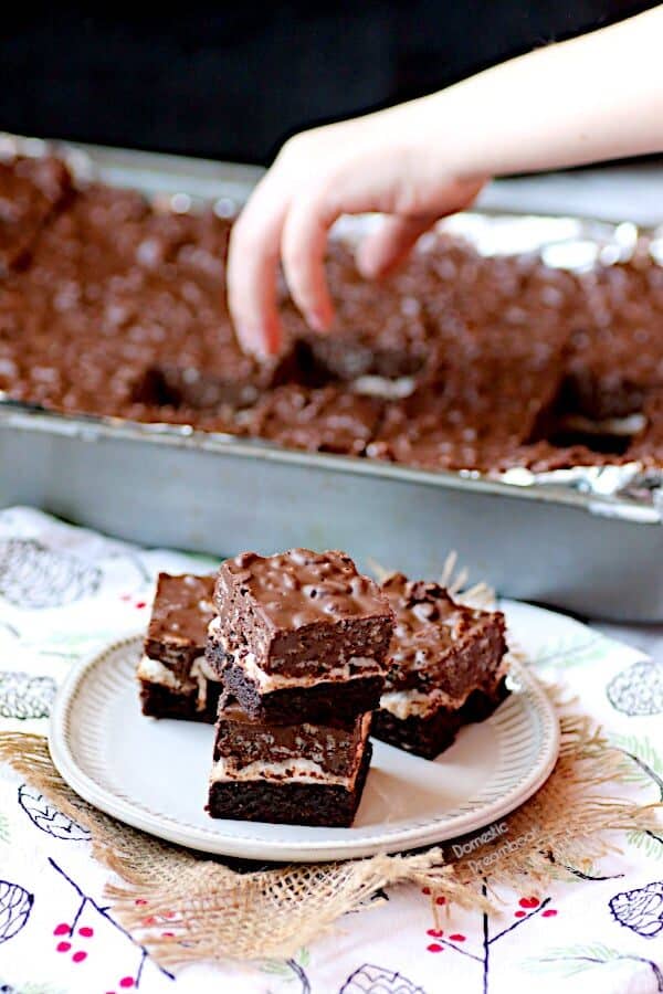A plate of chocolate peanut butter brownies with a hand grabbing a brownie from a pan in the background