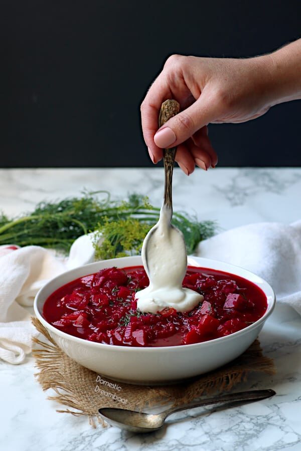 Beet borscht in a white bowl with a hand adding a dollop of sour cream.