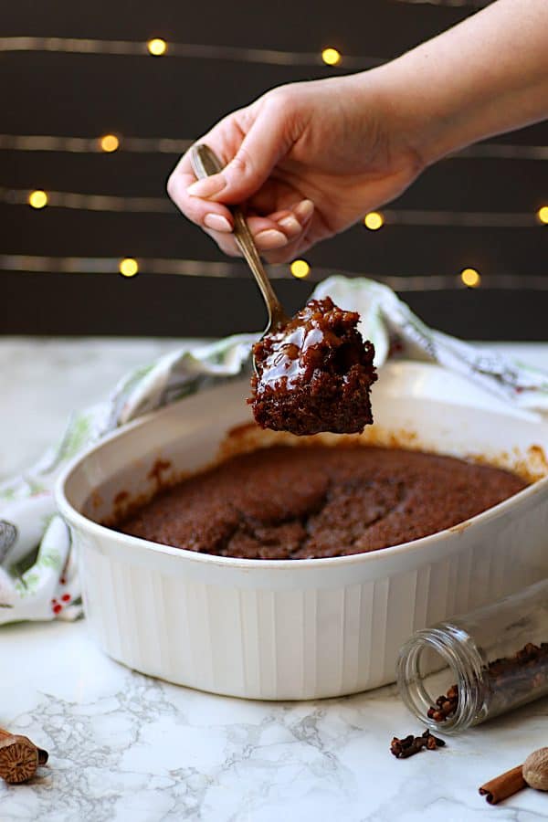 A hand scooping a spoonful of gingerbread pudding cake from a white casserole dish.