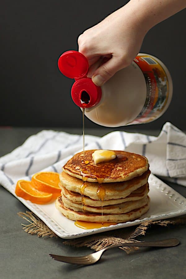 A hand pouring maple syrup on a stack of pancakes. 