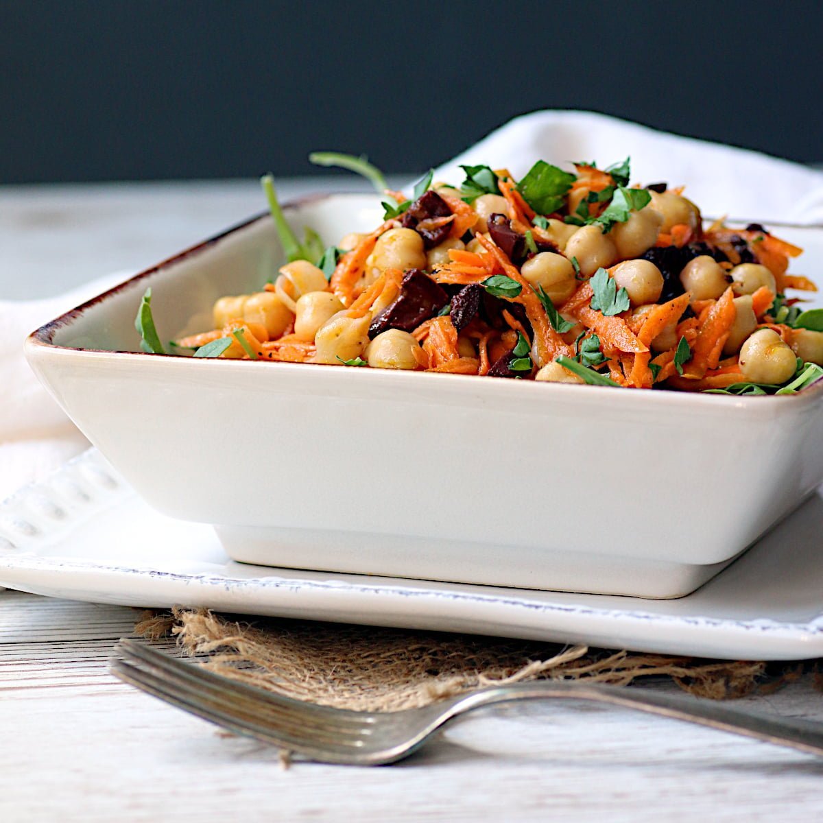Chickpea salad with carrots and olives in a square, white bowl.