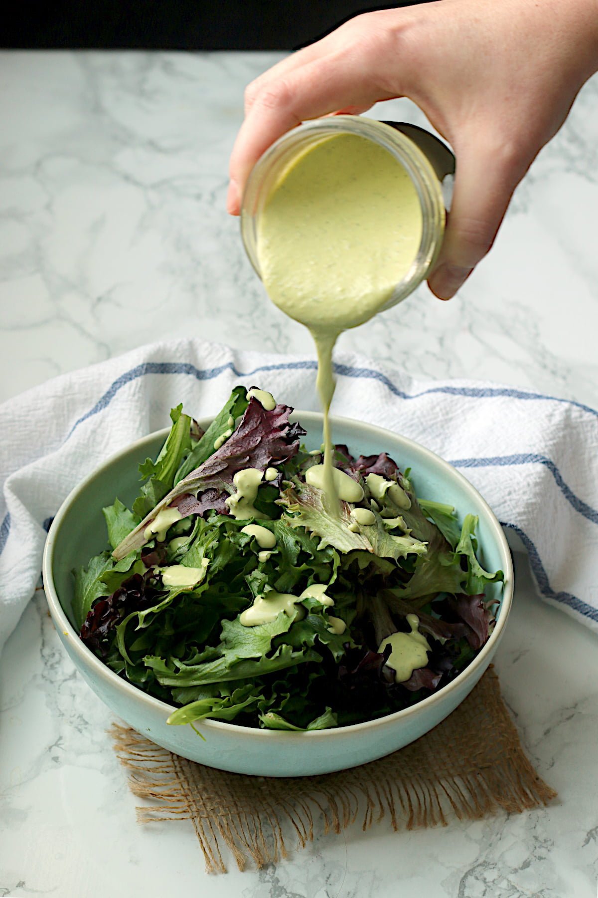 A hand pouring homemade creamy pesto salad dressing on a bowl of mixed salad greens.