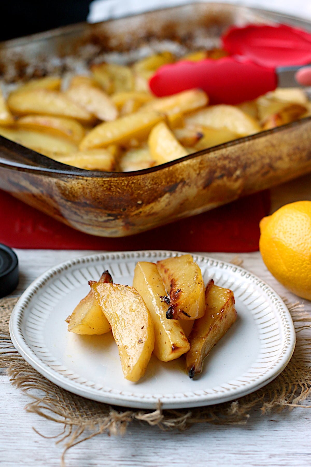 Greek Lemon Potatoes on a small white plate with a glass baking dish full of the same in the background.