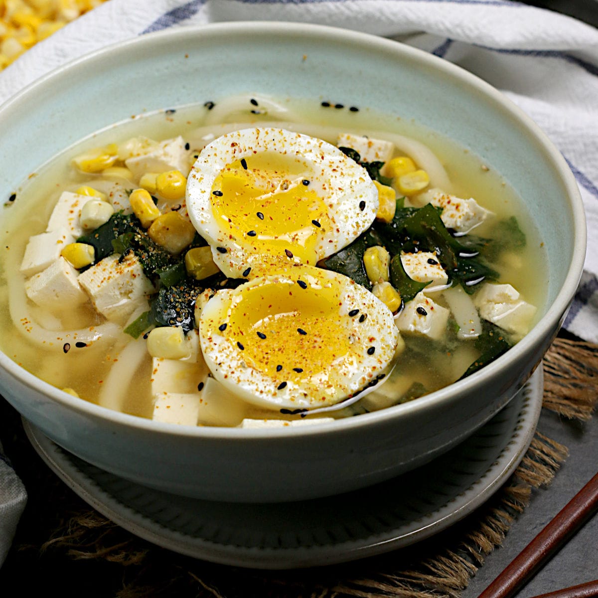 Miso Udon soup with wakame seaweed, corn, tofu, and a soft-boiled egg in a pale blue bowl.