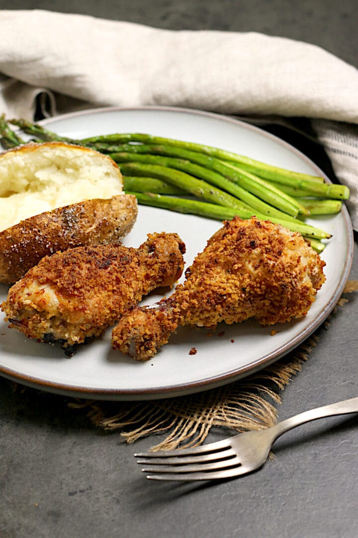 Perfect oven-fried chicken drumsticks with roasted asparagus and a baked potato on a white dinner plate.