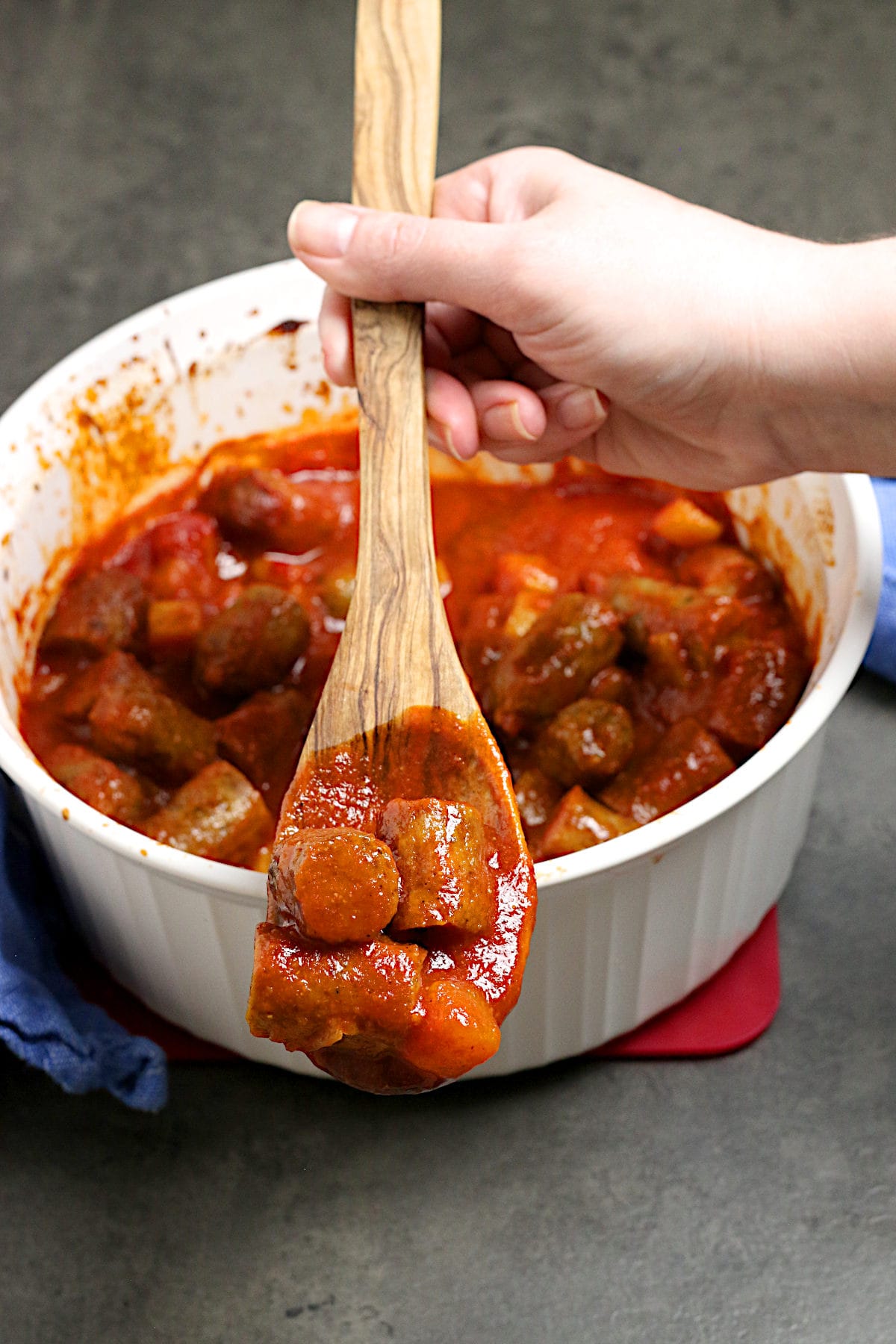 Peachy Sweet and Sour Sausages A hand scooping Peachy Sweet and Sour Sausages from a white casserole dish on a wooden spoon.