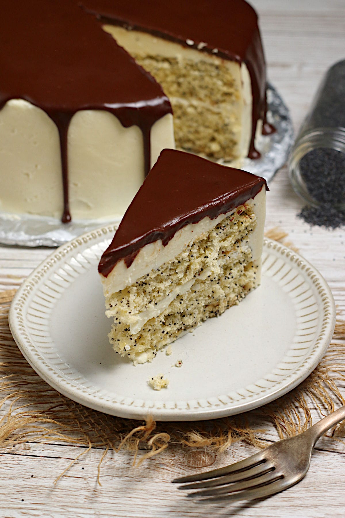 A slice of Poppyseed Sponge Cake with vanilla buttercream and chocolate ganache on a white plate.