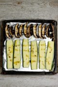 Sliced portabella mushrooms and zucchini drizzled with seasoned olive oil on a foil-lined baking pan.