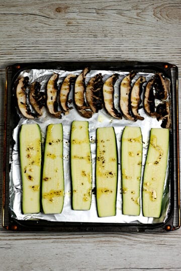 Sliced portabella mushrooms and zucchini drizzled with seasoned olive oil on a foil-lined baking pan.