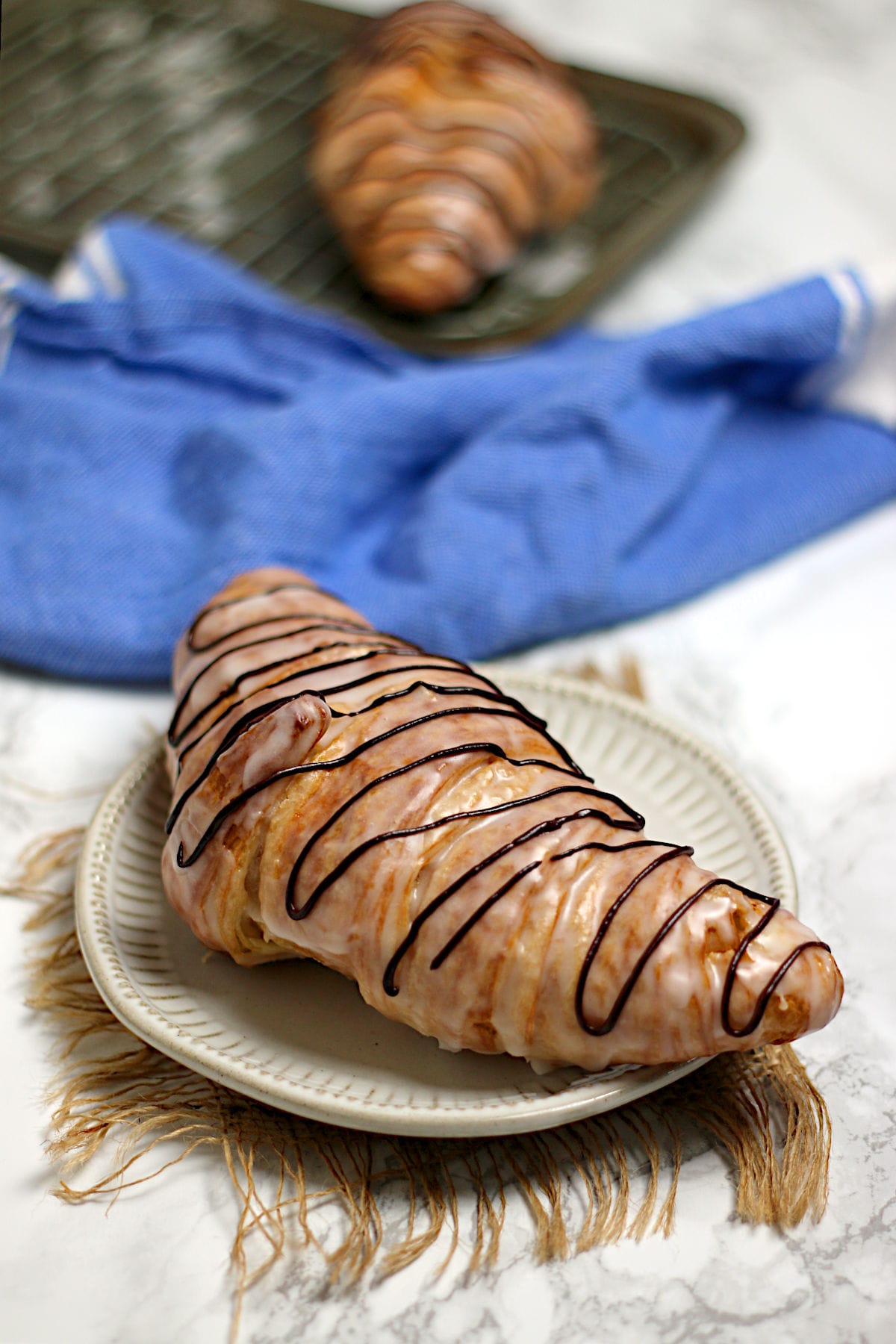 A chocolate drizzled glazed croissant on a small white plate.