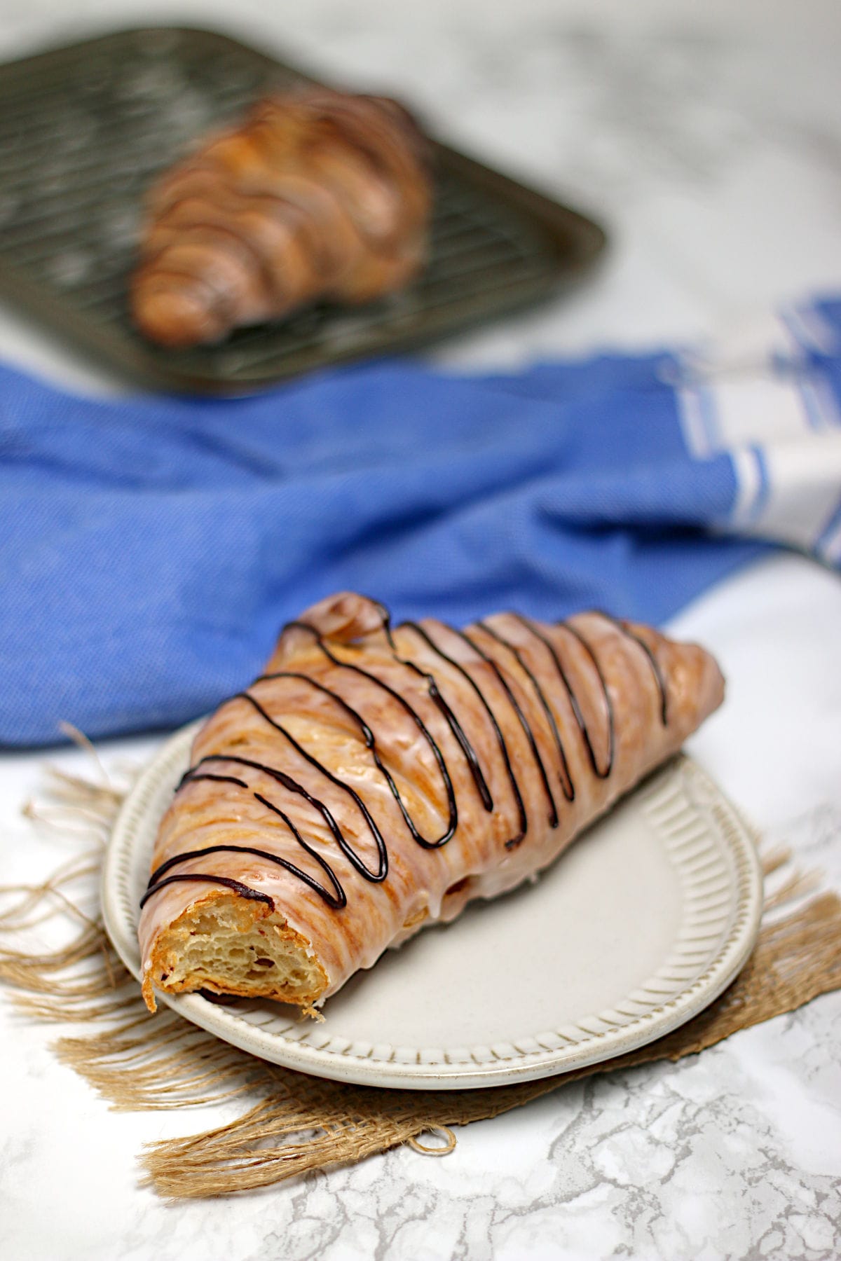 A chocolate drizzled glazed croissant with a bite missing on a small white plate.