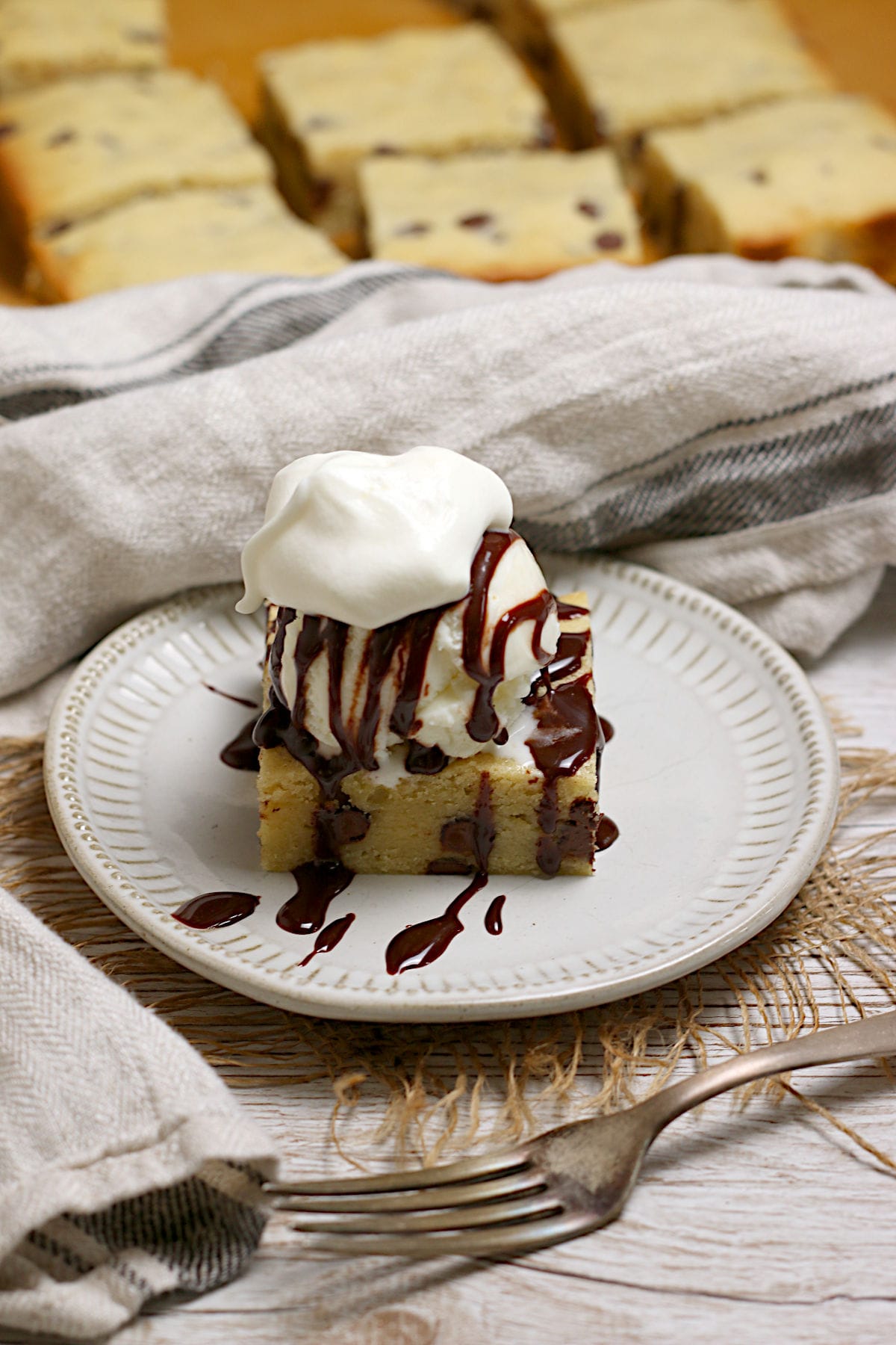 A white chocolate brownie topped with a scoop of vanilla ice cream, chocolate sauce, and a dollop of whipped cream on a small white plate.