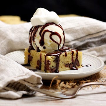 A white chocolate brownie topped with a scoop of vanilla ice cream, chocolate sauce, and a dollop of whipped cream on a small white plate.