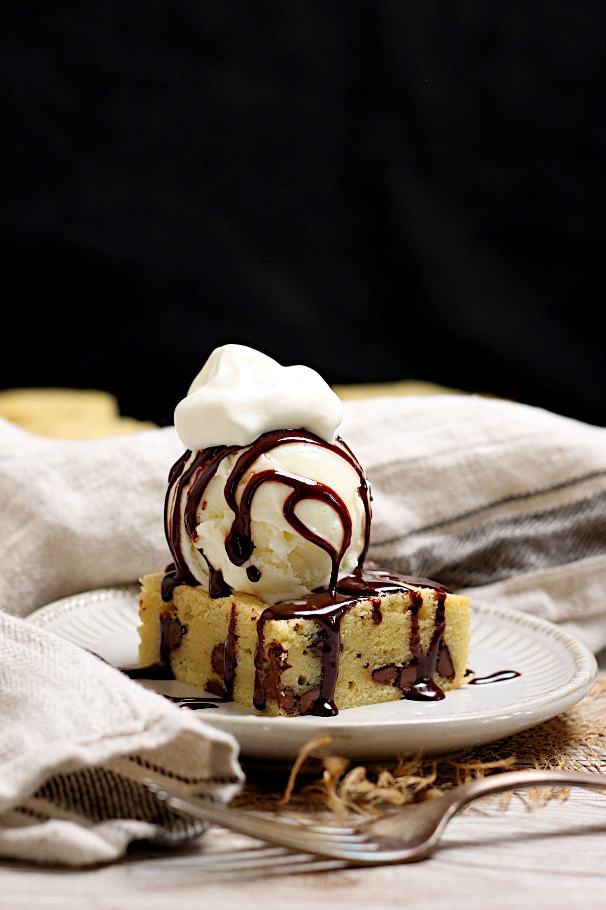 A white chocolate brownie topped with a scoop of vanilla ice cream, chocolate sauce, and a dollop of whipped cream on a small white plate.