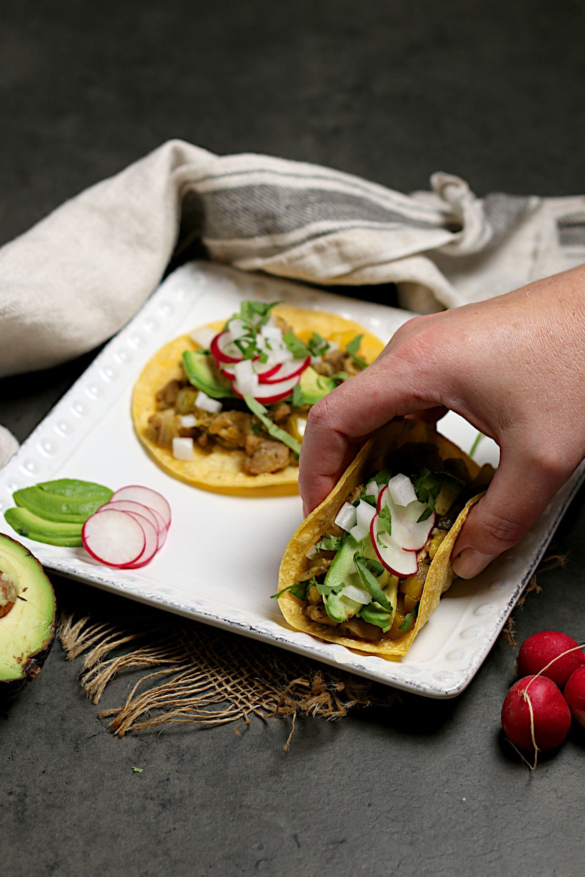 A hand picking up a Green Chile Chicken taco from a square white plate.