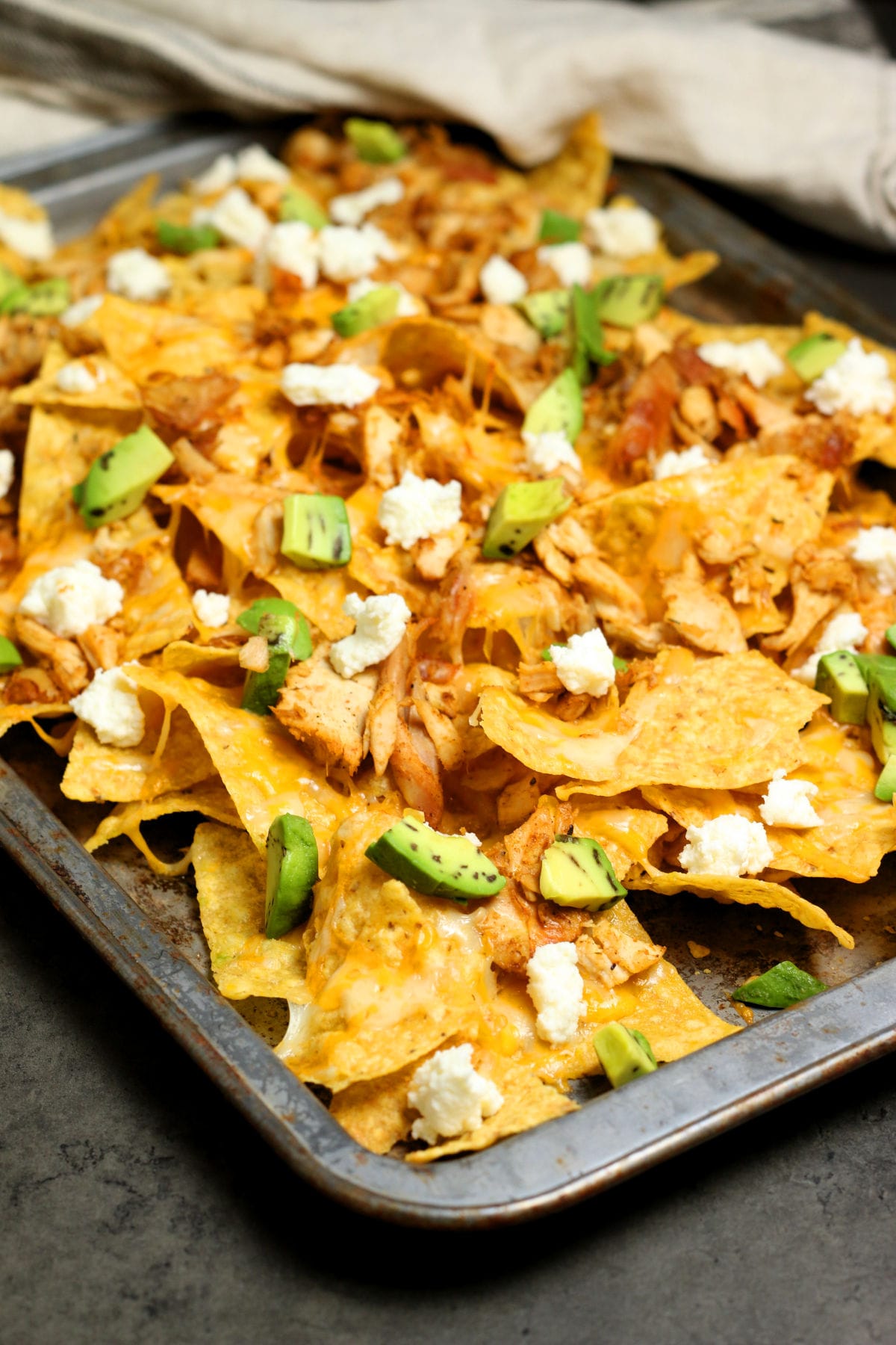 Homemade nachos on a baking sheet with cheddar cheese, chicken, avocado and homemade queso fresco.