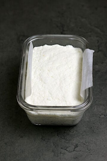 Homemade queso fresco pressed into a glass container lined with a strip of parchment paper.