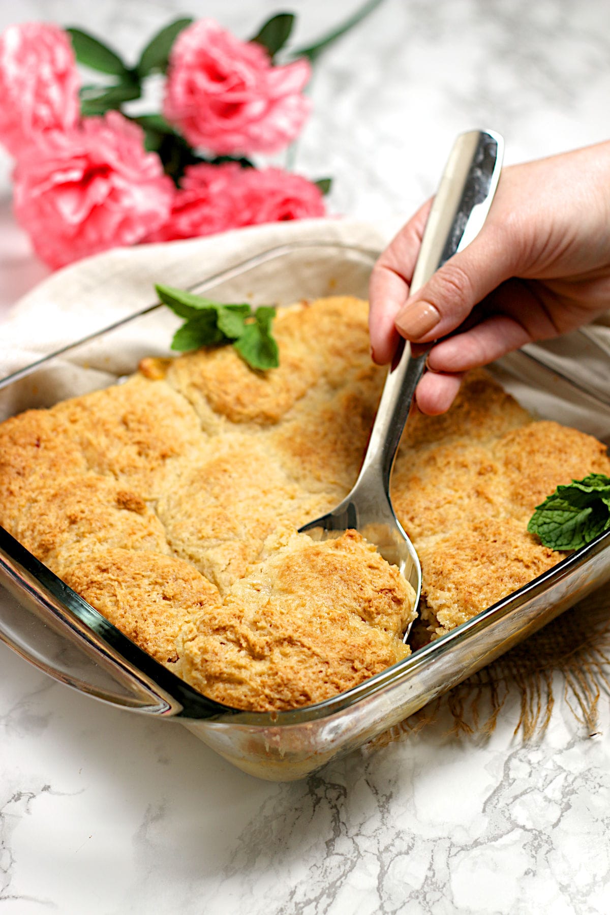 A hand scooping Peaches and Cream Cobbler from a glass baking dish.